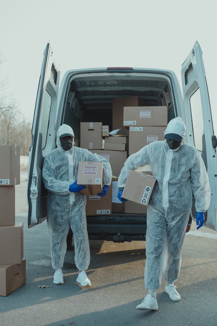 Logistics workers in protective gear unload packages from a delivery van outdoors.
